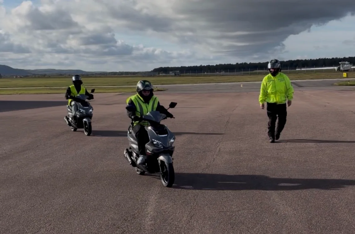 Två AM-elever kör moped medan läraren ger personlig handledning på ett stort och säkert övningsområde.