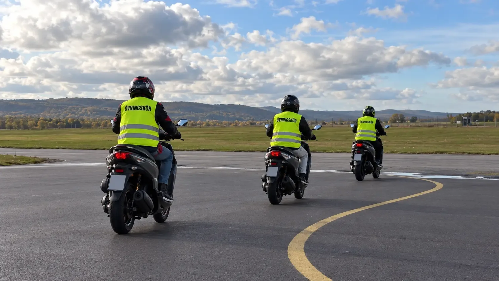 Tre AM-elever i reflexvästar med texten Övningskör kör moped på det stora övningsområdet.
